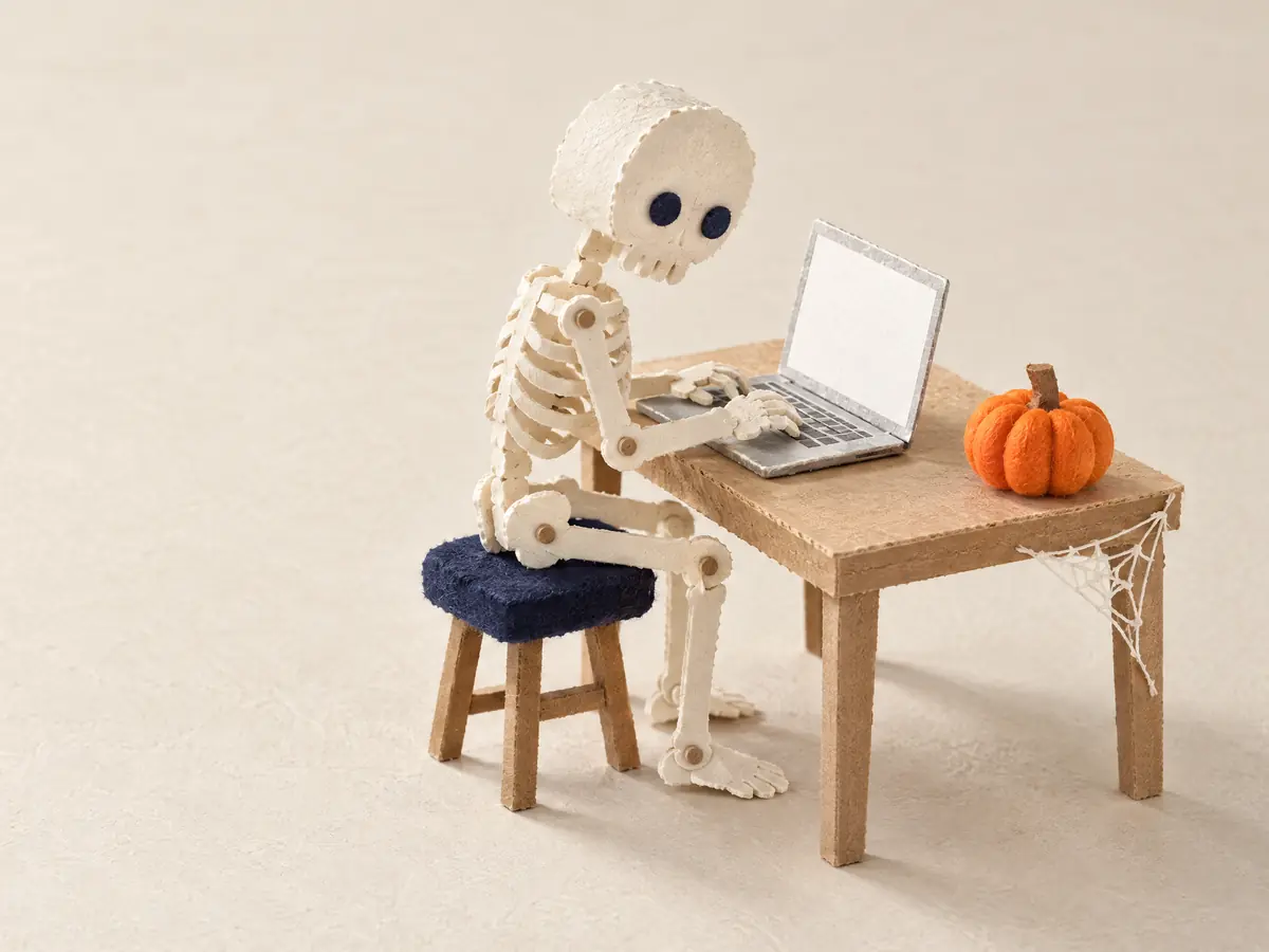 Een papieren skelet aan een klein bureau dat op een laptop typt, met een oranje pompoen ernaast