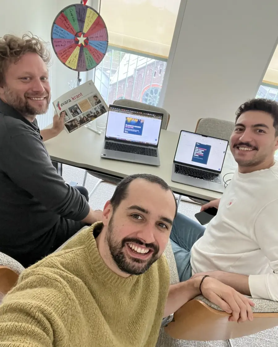 Three people smiling around a table with two open laptops showing post designs. One holds up a printed company newspaper, and a wheel covered in handwritten notes hangs behind them.