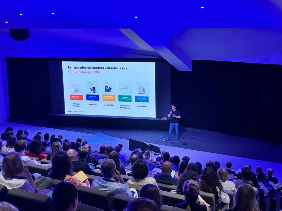 A speaker on stage in a full auditorium, in front of a slide in Dutch titled 'Een gevarieerde content kalender is key' with five content categories.